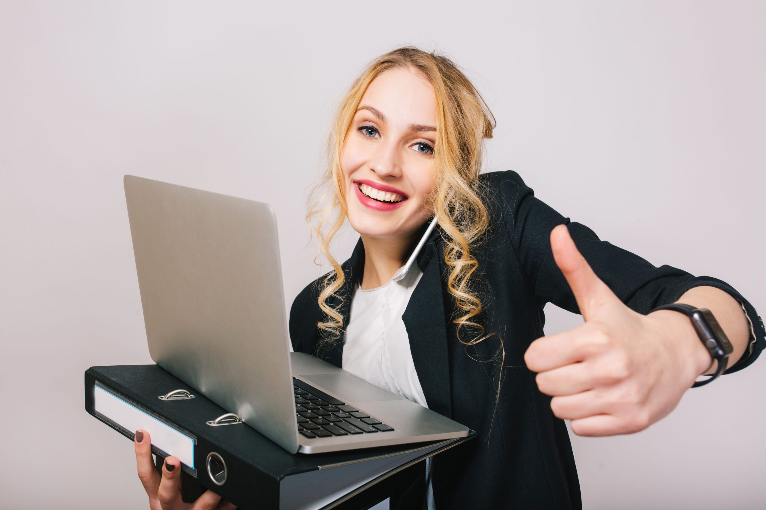 Portrait successful joyful businesswoman smiling to camera, holding laptop, folder, talking on phone isolated on white background. Wearing white shirt and black jacket, modern office worker.