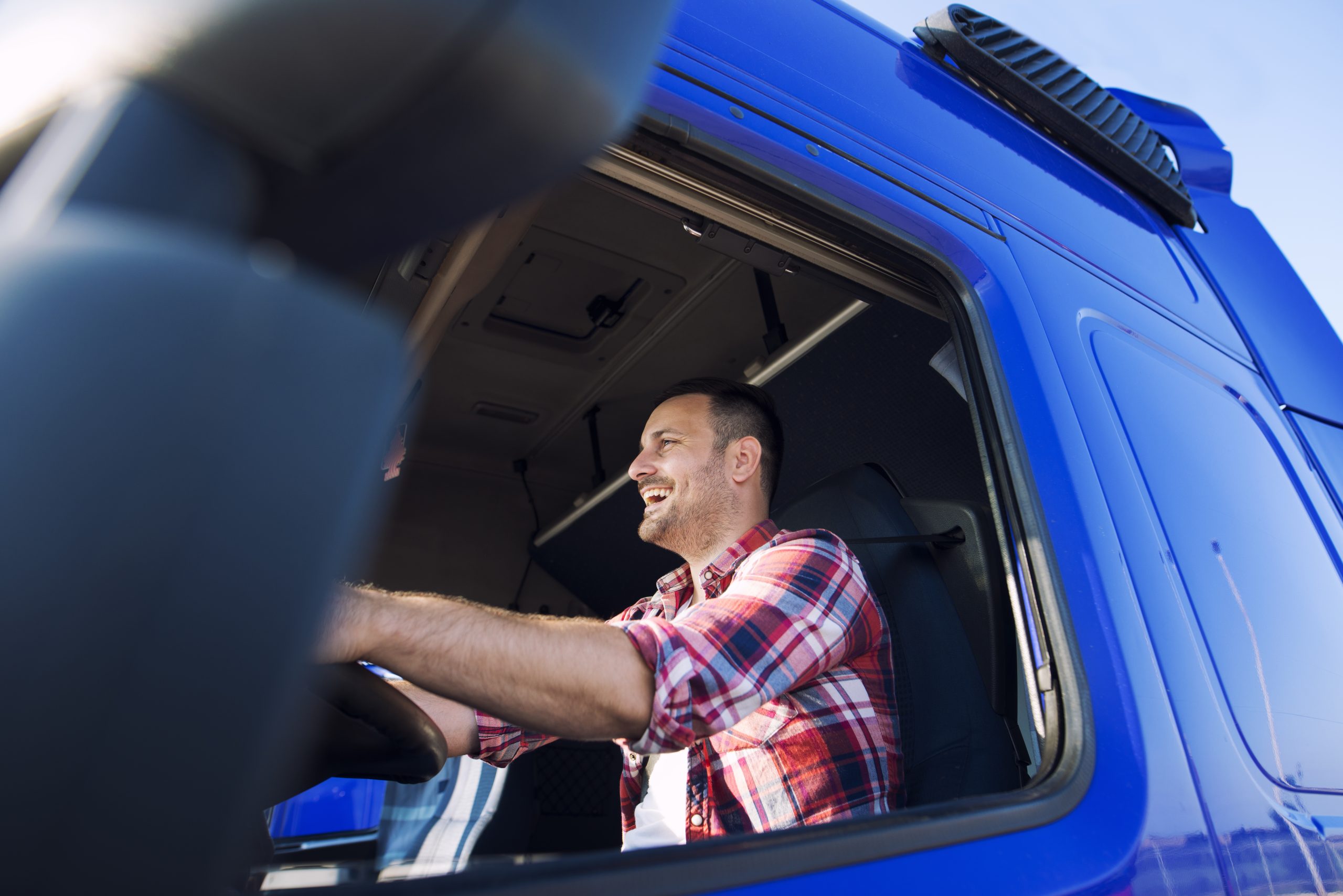 Professional middle aged trucker in cabin driving truck and smiling.